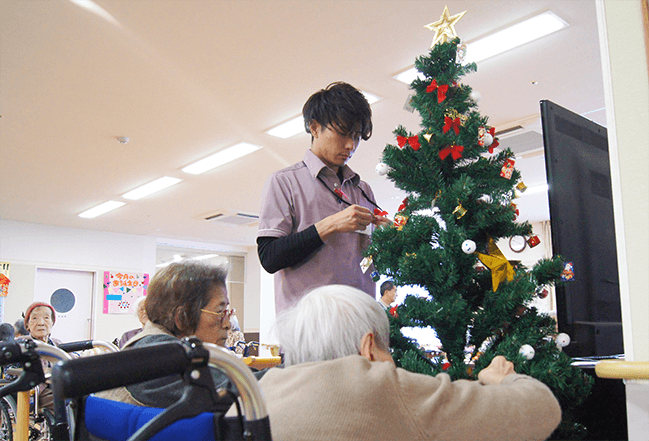 介護施設で介護職員と利用者が一緒にクリスマスツリーの飾りつけをしている